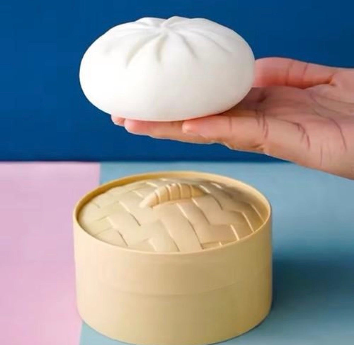 Hand holding a white dumpling squishy above its open bamboo-style steamer